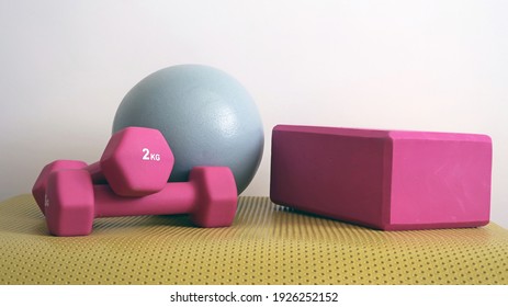 Home exercise equipment. A pair of pink dumb bells, resting on a yoga mat, a small grey Pilates stability ball and a styrofoam yoga block or brick. Light grey background wall. Close up
