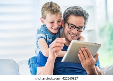 At home, a father and his young son having fun by gaming on a tablet, dad sits on a white couch and the boy looks at the screen over the shoulder of his father and giving him advices to win - Powered by Shutterstock