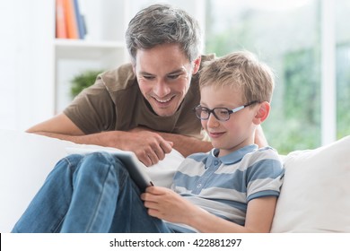 At Home, A Father And His Young Son Having Fun By Gaming On A Tablet, Kid Sits On A White Couch And Dad Looks At The Screen Over The Shoulder Of His Son And Giving Him Advices To Win