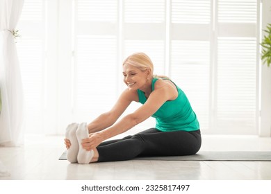 Home Fitness. Portrait Of Beautiful Senior Woman Stretching Leg Muscles, Sporty Mature Lady Sitting On Fitness Mat In Light Living Room, Enjoying Domestic Trainings And Healthy Lifestyle, Copy Space - Powered by Shutterstock