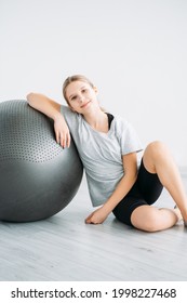 Home Fitness Training. Kid Sport. Pilates Equipment. Physio Therapy Accessories. Smiling Relaxed Girl Sitting On Floor With Exercise Ball On Light Background.