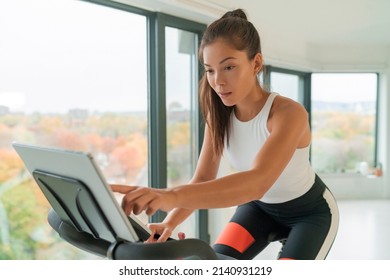 Home fitness workout woman training on smart stationary bike indoors watching screen connected online to live streaming subscription service for biking exercise. Young Asian woman athlete.