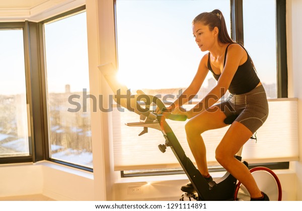 Home fitness workout woman training on smart\
stationary bike indoors watching screen connected online to live\
streaming subscription service for biking exercise. Young Asian\
woman athlete.