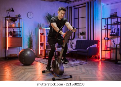 Home Fitness Workout, Young Asian Man Athlete Training On Smart Stationary Bike Indoors Watching On Screen Connected Online To Live Streaming Subscription Service For Biking Exercise.