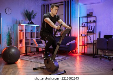 Home Fitness Workout, Young Asian Man Athlete Training On Smart Stationary Bike Indoors Watching On Screen Connected Online To Live Streaming Subscription Service For Biking Exercise.