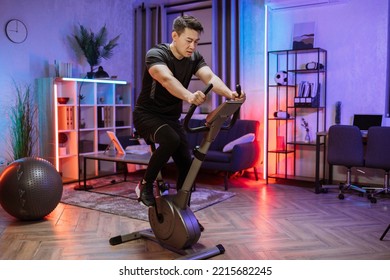 Home Fitness Workout, Young Asian Man Athlete Training On Smart Stationary Bike Indoors Watching On Screen Connected Online To Live Streaming Subscription Service For Biking Exercise.