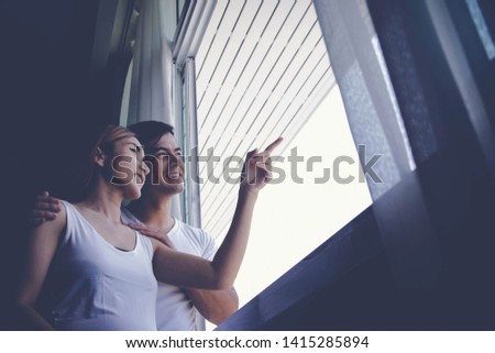 Home Happiness Concept : The couple, husband and wife are pregnant, standing by the window, the wife pointing to the husband looking at what she pointed out outside happily.

