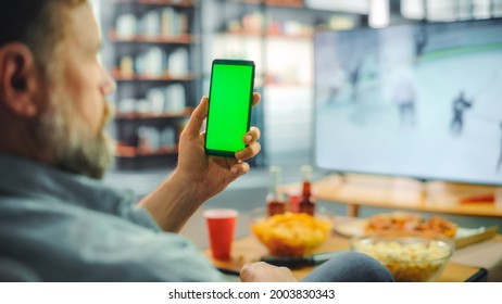 At Home Ice Hockey Fan Holds Green Screen Chroma Key Smartphone, Friends Watch Game On TV, Cheer For Favourtite Sports Team To Win Championship. Professional Club Play. Over The Shoulder Close-up