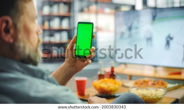 At Home Ice Hockey Fan Holds Green Screen Chroma\
Key Smartphone, Friends Watch Game on TV, Cheer for Favourtite\
Sports Team to Win Championship. Professional Club Play. Over the\
Shoulder Close-up