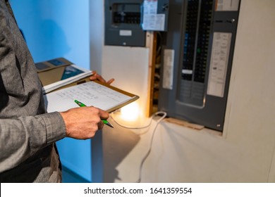 Home Inspector In Front Of Electric Distribution Board During Inspection, Selective Focus And Close Up On Man's Hands As He Holding Notebook And Pen