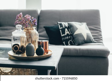 Home interior decor in gray and brown colors: glass jar with dried flowers, vase and candle on the wooden tray on the coffee table over sofa with cushions. Living room decoration. - Shutterstock ID 715596850