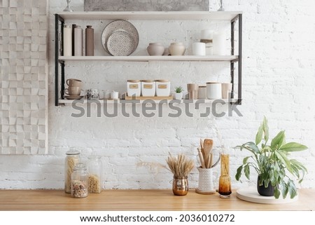 Home interior decoration, closeup of white kitchen. Modern decor of scandinavian style house, wooden shelf with decorative ceramic tableware. Rustic glass details at tabletop furniture.