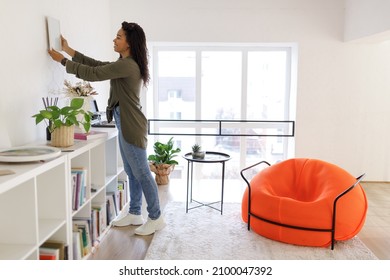 Home Interior And Domestic Decor. Smiling Young Black Woman Hanging Painting, Putting Photo Picture Frame On The Wall. Casual Interior Designer Taking Care Of Coziness In New Modern Stylish Apartment