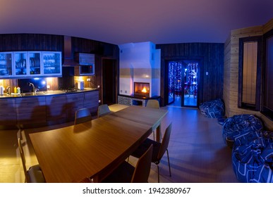 Home Interior Kitchen. Large Wooden Table Is Located In Kitchen. Evening Interior With Neon Lighting. Dining Room With Fireplace. Rest Room Is Combined With Kitchen. Fireplace In Lounge.