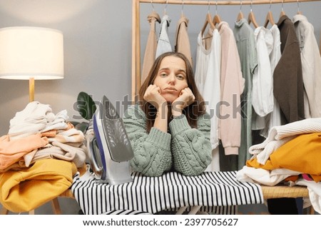 Home ironing routine. Household wardrobe care. Bored sad adult woman wearing casual shirt ironing clothing doing household chores after laundry having lots of work