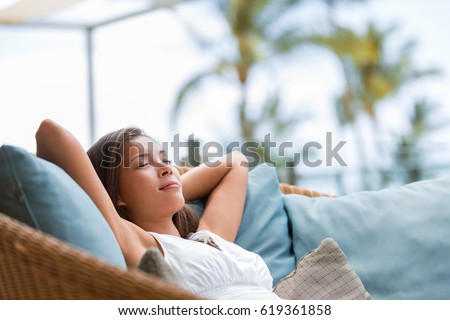Home lifestyle woman relaxing sleeping on sofa on outdoor patio living room. Happy lady lying down on comfortable pillows taking a nap for wellness and health. Tropical vacation.