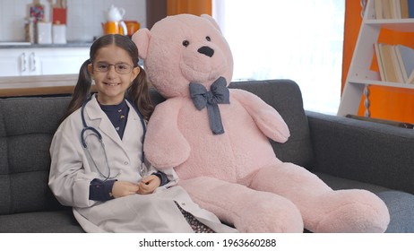 At Home, The Little Girl Wearing A Doctor's Apron With Glasses Looks At Her Teddy Bear, Corrects Her Glasses And Poses With The Bear. Portrait Of Child Giving Doctor Pose With Teddy Bear.