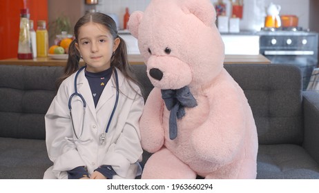 At Home, The Little Girl Wearing A Doctor's Apron With Glasses Looks At Her Teddy Bear, Corrects Her Glasses And Poses With The Bear. Portrait Of Child Giving Doctor Pose With Teddy Bear.