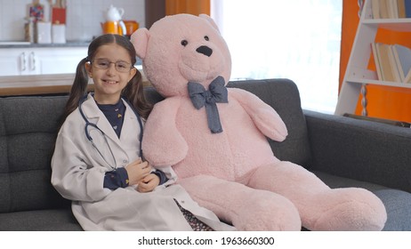 At Home, The Little Girl Wearing A Doctor's Apron With Glasses Looks At Her Teddy Bear, Corrects Her Glasses And Poses With The Bear. Portrait Of Child Giving Doctor Pose With Teddy Bear.