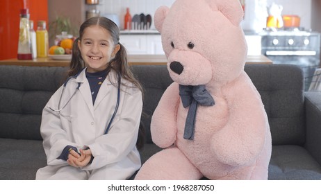 At Home, The Little Girl Wearing A Doctor's Apron With Glasses Looks At Her Teddy Bear, Corrects Her Glasses And Poses With The Bear. Portrait Of Child Giving Doctor Pose With Teddy Bear.