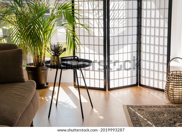 Home living room with wood and paper divider\
screen blocking sun from window, black metal accent table and sofa,\
natural colors.