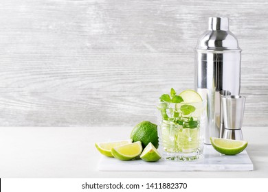 Home Made Mojito Cocktail Decorated With Lime, Mint Leaves, Ice Cubes And Silver Shaker Over White Bright Background. Copy Space