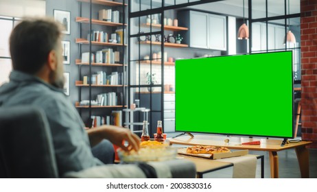 At Home Man Sitting On A Couch Watch Green Screen Chroma Key TV. Middle Aged Male Resting, Watches Show, Sports Event, Film, On Demand Streaming Service, Eats Snacks. Over The Shoulder Shot