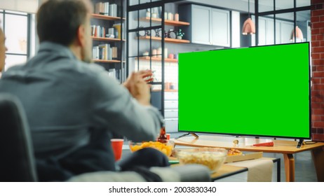 At Home Man Sitting On A Couch Watch Green Screen Chroma Key TV. Middle Aged Male Resting, Watches Show, Sports Event, Film, On Demand Streaming Service, Eats Snacks. Over The Shoulder Shot