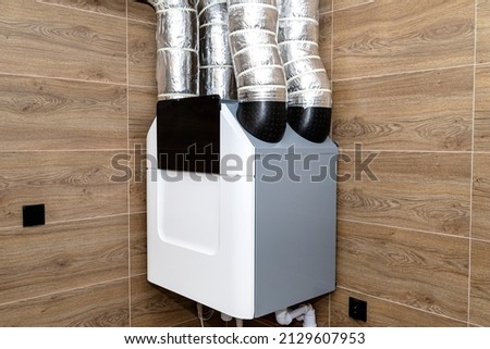 Home mechanical ventilation with heat recovery hanging on the wall in a modern gas boiler room with brown ceramic tiles imitating wood.
