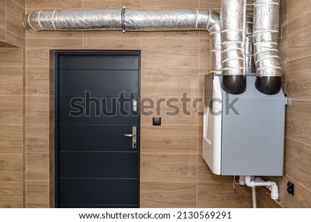 Home mechanical ventilation with heat recovery hanging on the wall in a modern gas boiler room with brown ceramic tiles imitating wood.