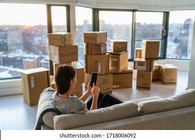 Home move out of apartment moving boxes woman using online movers services on mobile phone app easy pick-up with packages for new home. Asian new homeowner girl happy sitting in sofa.