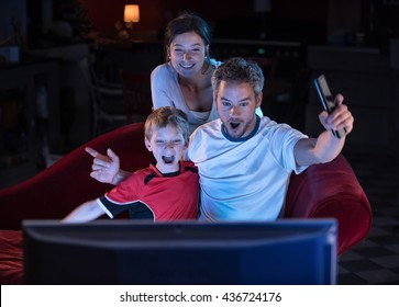 At Home, Night Falls, A Nice Family Of Three People Watching Sport At  TV, They Raise Their Arms In Victory, Daddy Has The Remote In Hand