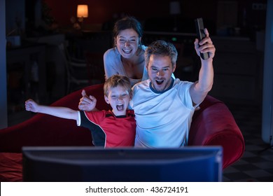 At Home, Night Falls, A Nice Family Of Three People Watching Sport At  TV, They Raise Their Arms In Victory, Daddy Has The Remote In Hand