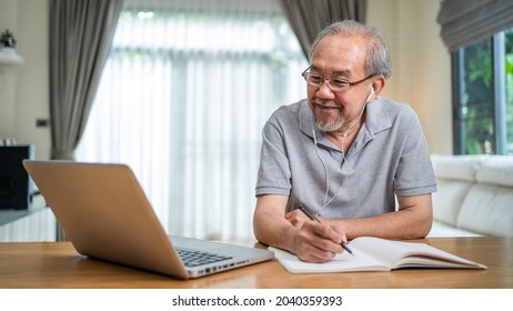 Home Office Of Asian Senior Elderly Business Man Working Online With Laptop Computer. Mature Older Male Talking In Online Conference Video Call And Write In Copy Book In House During COVID-19 Pandemic