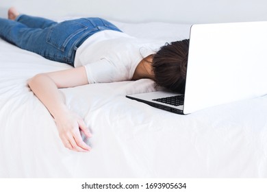 Home Office In Bed. Young Woman Wearing White Shirt And Blue Jeans And Sleeping On A White Laptop In Bed With White Sheets