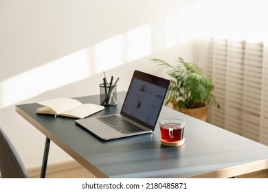 Home Office Concept. Designated Work From Home Area Near The Window. Modern Laptop With Blurred Screen And Glass Cup Of Tea On Table. Close Up, Copy Space, Interior Background.