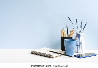 Home And Office Desk With Notepad, Phone, Pencils, Coffee, On A White Table Over Light Blue Wall.