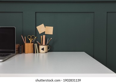 Home Office With Laptop In The Corner, Office Supplies, Mug With Tools, Notes, Books And Dark Green Wall. Homeschooling Concept. Trendy, Creative Workspace.