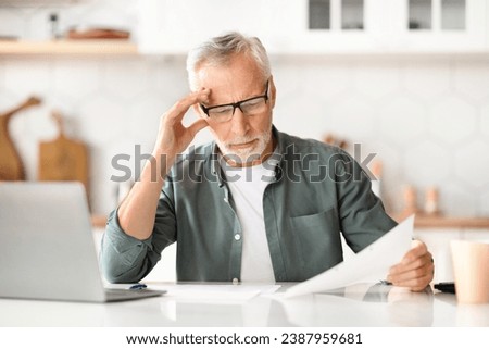 Home Office Paperwork. Portrait Of Senior Gentleman At Kitchen Desk, Reviewing Documents And Using Laptop, Elderly Man Engaged In Remote Work, Reading Financial Papers At Home, Free Space