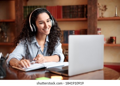 Home Office. Portrait of smiling young lady in wireless headphones sitting at desk, writing in notebook and using computer. Woman watching online tutorial, webinar or seminar, studying at home