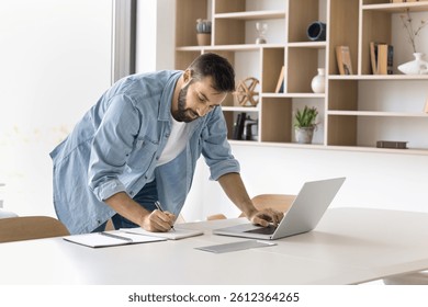 Home office workflow. Focused millennial man studying or working on distance stand by workplace table browse internet for information write notes in paper copybook managing tasks planning work process