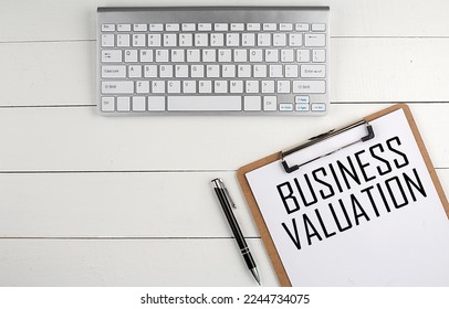 Home office workspace with keyboard, clipboard and pen with text BUSINESS VALUATION on a white wooden background , business concept