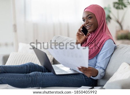 Home Office. Young Black Muslim Freelancer Woman Working With Papers And Laptop In Living Room And Talking On Cellphone, African Islamic Lady In Hijab Sitting On Couch Enjoying Remote Job, Copy Space
