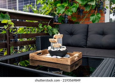 Home patio with black plastic garden furniture, small relaxing electrical zen table fountain on table and real grape vines with grapes hanging on background. Home decor.