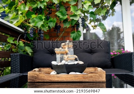 Home patio with black plastic garden furniture, small relaxing electrical zen table fountain on table and real grape vines with grapes hanging on background. Home decor.