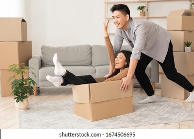 Home, People, Moving And Real Estate Concept. Excited Asian Couple Having Fun And Riding In Cardboard Boxes In New Flat