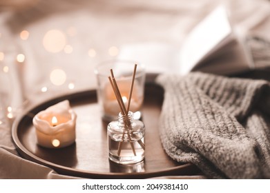 Home perfume in glass bottle with wood sticks, scented burn candles, open paper book and knit wool textile on ray in bedroom close up. Aromatherapy cozy atmosphere lifestyle. Winter warm xmas season.  - Shutterstock ID 2049381410
