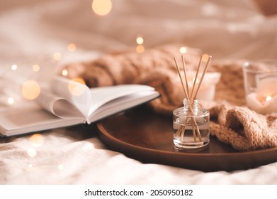 Home perfume in glass bottle with wood sticks, scented burn candles, open paper book and knit wool textile on ray in bedroom close up. Aromatherapy cozy atmosphere lifestyle. Winter warm xmas season.  - Shutterstock ID 2050950182