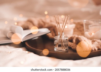 Home Perfume In Glass Bottle With Wood Sticks, Scented Burn Candles, Open Paper Book And Knit Wool Textile On Ray In Bedroom Close Up. Aromatherapy Cozy Atmosphere Lifestyle. Winter Warm Xmas Season. 