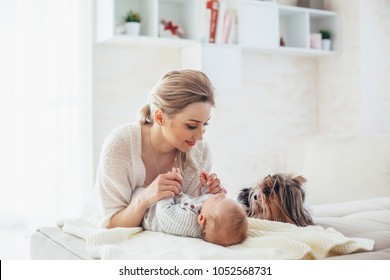 Home Portrait Of A 2 Month Old Baby With Mom And Dog On The Bed. Mother Playing With The Child And Looking To A Pet.